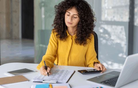 Woman in a yellow blouse using a calculator while reviewing a financial spreadsheet at her desk.