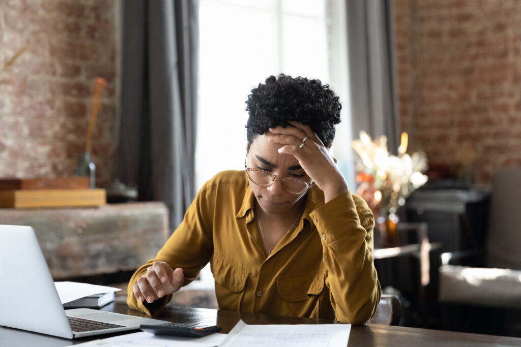 Person sitting at a desk with a laptop and calculator, appearing stressed while reviewing financial documents.