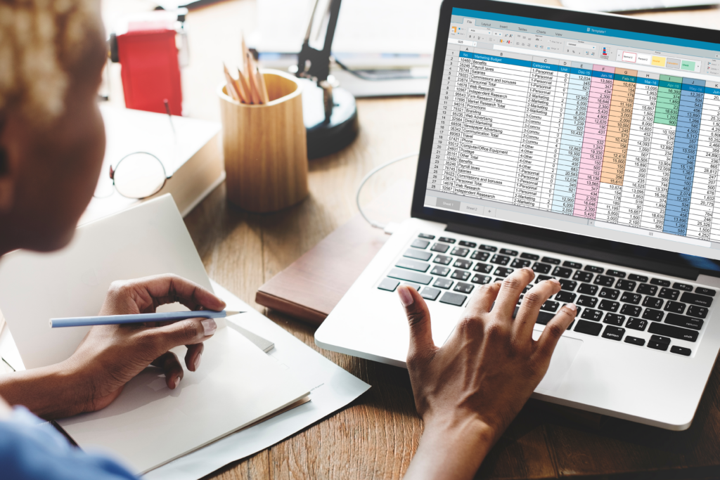 Person working at a desk with a laptop displaying a colorful spreadsheet and taking notes in a notebook.