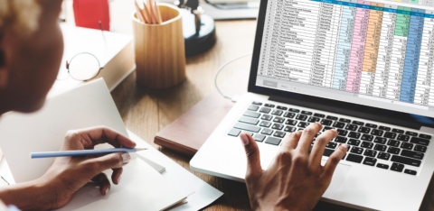 Person working at a desk with a laptop displaying a colorful spreadsheet and taking notes in a notebook.