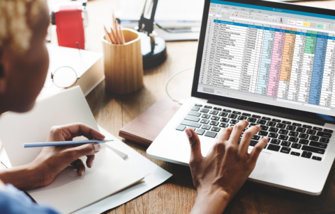 Person working at a desk with a laptop displaying a colorful spreadsheet and taking notes in a notebook.