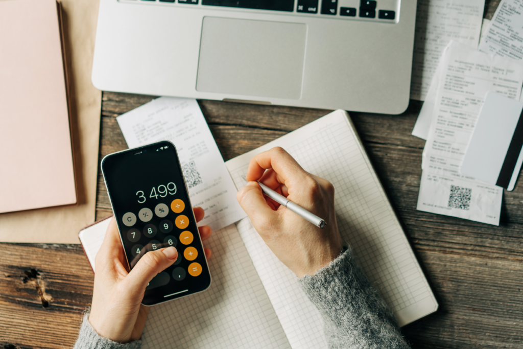 Person using a smartphone calculator and writing in a notebook with receipts and a laptop on a wooden table.