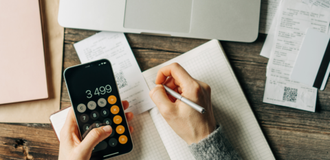 Person using a smartphone calculator and writing in a notebook with receipts and a laptop on a wooden table.