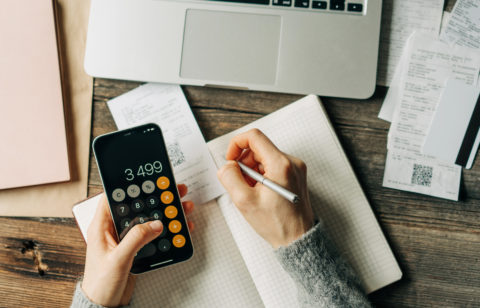 Person using a smartphone calculator and writing in a notebook with receipts and a laptop on a wooden table.