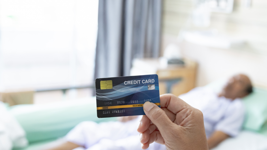 A person holding a credit card in the foreground while a patient lies in a hospital bed in the background.