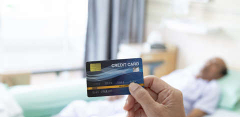 A person holding a credit card in the foreground while a patient lies in a hospital bed in the background.