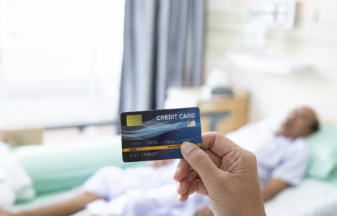A person holding a credit card in the foreground while a patient lies in a hospital bed in the background.