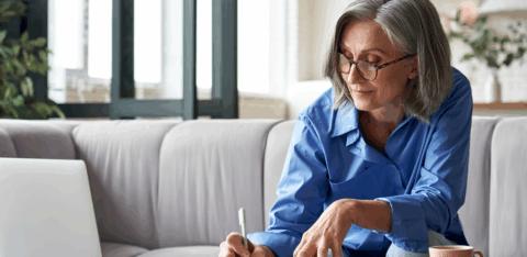 Older woman with gray hair and glasses sitting on a couch, reviewing paperwork and writing in a notebook while using a laptop at home.