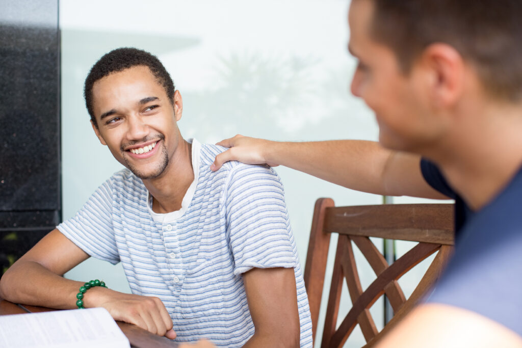 Smiling young man sitting at a table while another person offers a supportive hand on his shoulder.