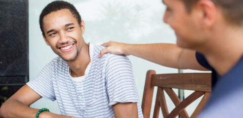 Smiling young man sitting at a table while another person offers a supportive hand on his shoulder.
