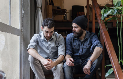 Two young men sitting on stairs indoors, one looking down with folded hands while the other looks at him and speaks.