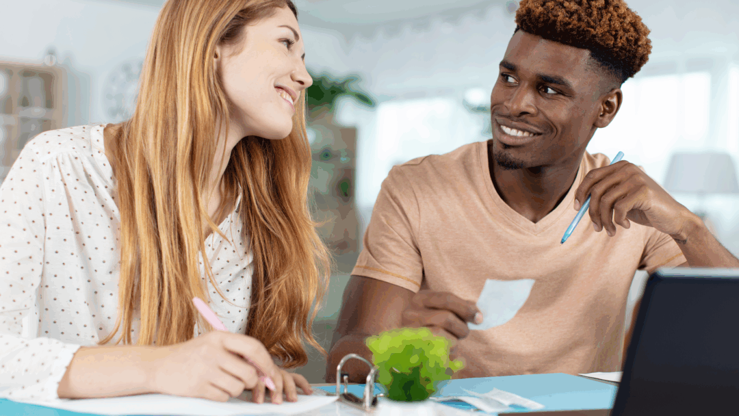Smiling couple working together at a table with papers, receipts, and a laptop, possibly budgeting or reviewing finances.