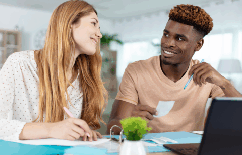 Smiling couple working together at a table with papers, receipts, and a laptop, possibly budgeting or reviewing finances.