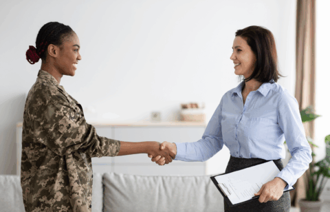 Smiling woman in military uniform shaking hands with a professional woman holding a clipboard.