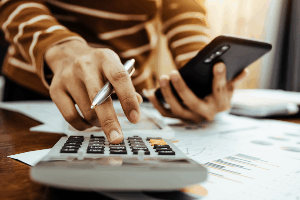 Close-up of a person using a calculator and holding a smartphone while reviewing printed financial documents.