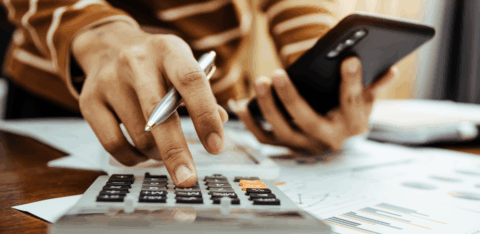 Close-up of a person using a calculator and holding a smartphone while reviewing printed financial documents.
