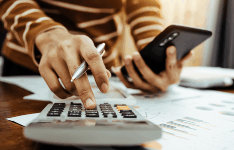 Close-up of a person using a calculator and holding a smartphone while reviewing printed financial documents.