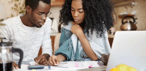 Couple sitting at a kitchen table, reviewing financial papers and using a calculator, with a laptop and fruit in the foreground.