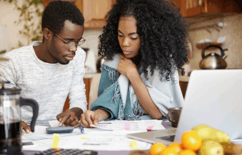 Couple sitting at a kitchen table, reviewing financial papers and using a calculator, with a laptop and fruit in the foreground.
