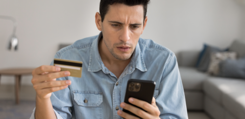 Man holding a credit card and looking concerned while checking his smartphone.