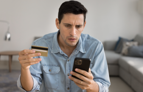 Man holding a credit card and looking concerned while checking his smartphone.