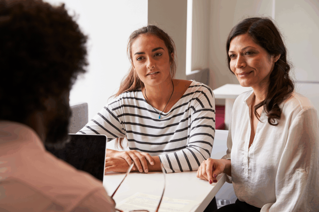Two women sit across a table from a man during a meeting, listening attentively and smiling.