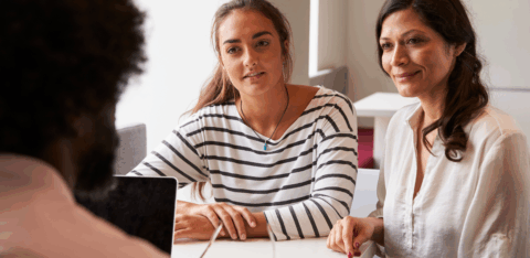 Two women sit across a table from a man during a meeting, listening attentively and smiling.