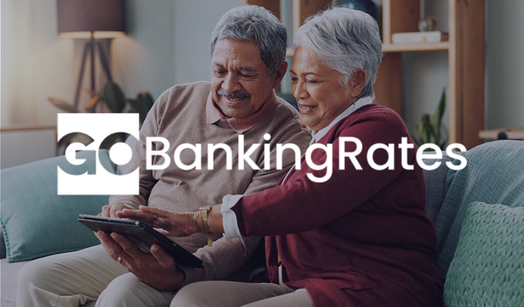 Smiling older couple enjoying time together on a couch while looking at a tablet, with the GOBankingRates logo overlaid.