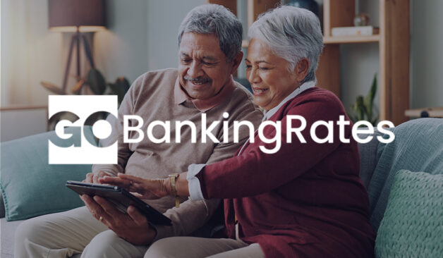 Smiling older couple enjoying time together on a couch while looking at a tablet, with the GOBankingRates logo overlaid.