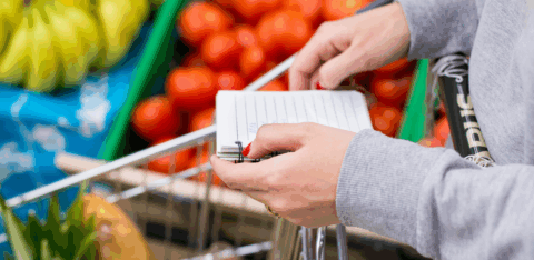 Person holding a shopping list while pushing a grocery cart filled with fresh produce in a supermarket.