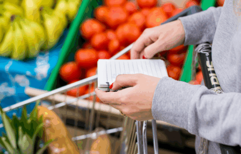 Person holding a shopping list while pushing a grocery cart filled with fresh produce in a supermarket.