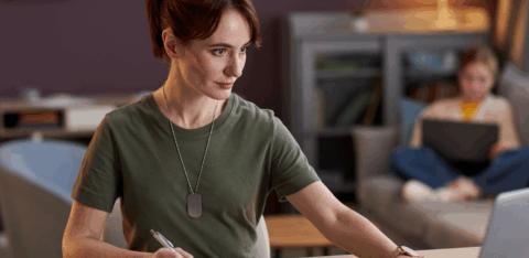 Veteran working on financial planning at a desk, with papers, a laptop, and another person in the background using a computer.