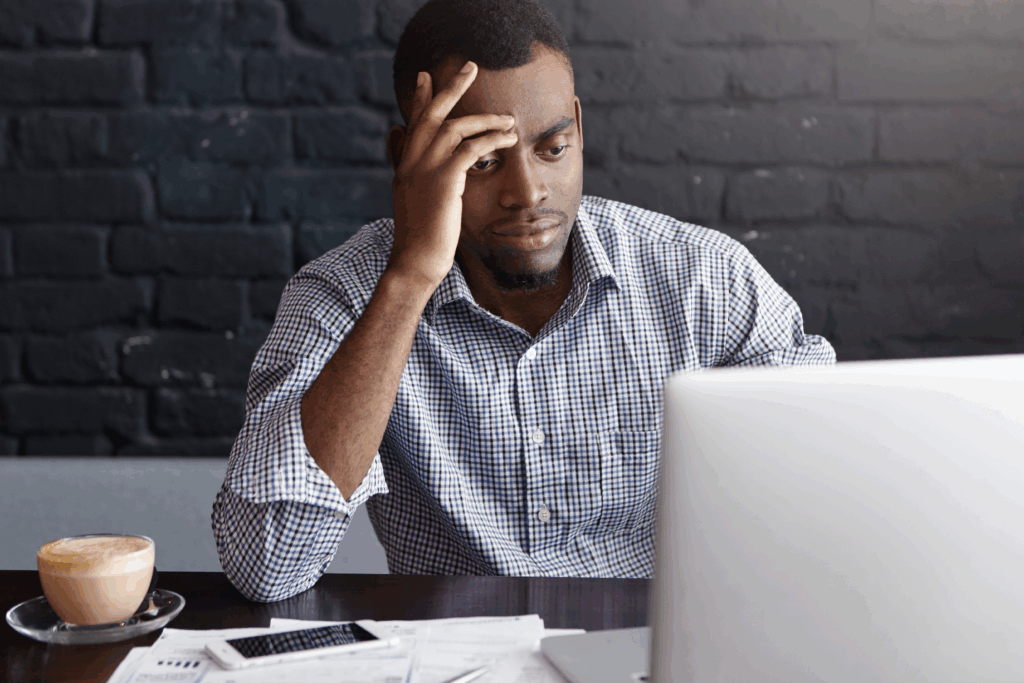 Man sitting at a table with a laptop, looking stressed while reviewing financial papers and a phone beside him.