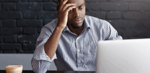 Man sitting at a table with a laptop, looking stressed while reviewing financial papers and a phone beside him.