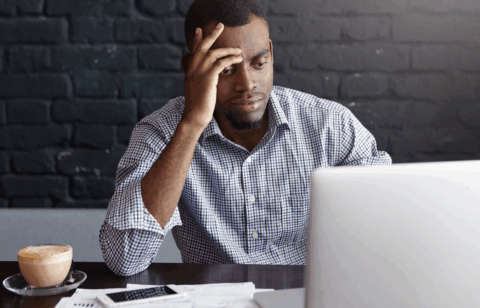 Man sitting at a table with a laptop, looking stressed while reviewing financial papers and a phone beside him.