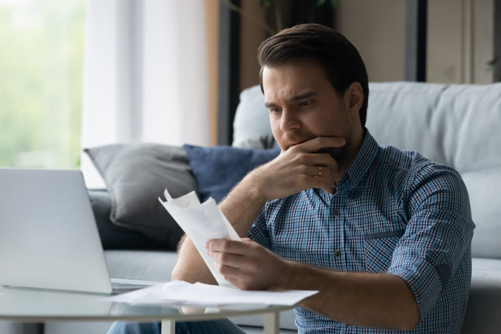 Man sitting on a couch, looking at bills with a concerned expression while using a laptop.