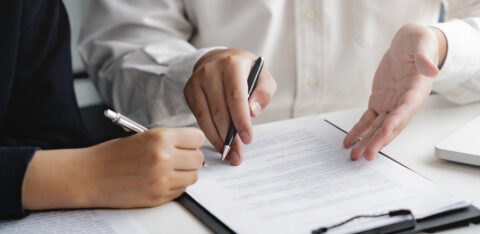 lose-up of two people reviewing and signing a contract on a clipboard at a desk.