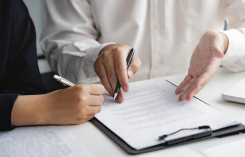 lose-up of two people reviewing and signing a contract on a clipboard at a desk.