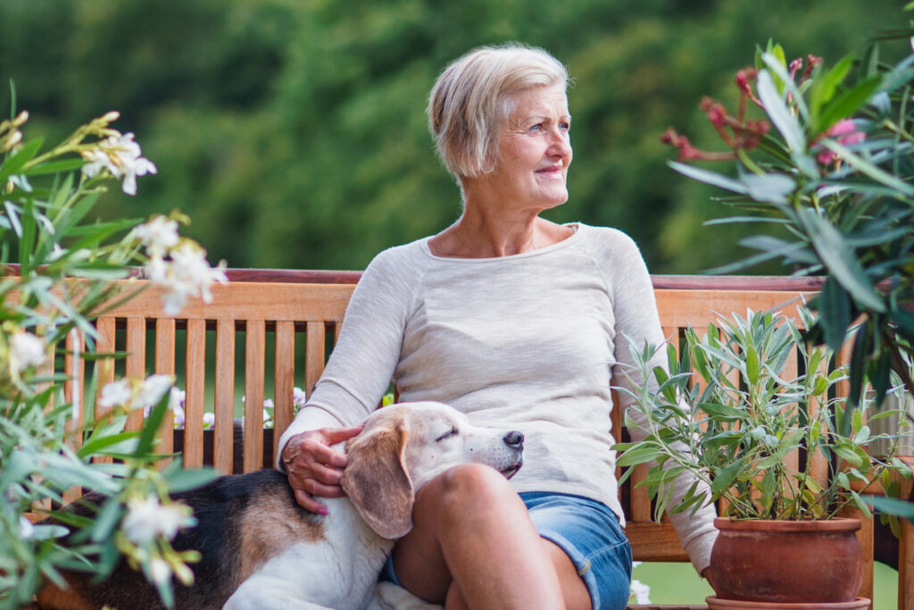 Older woman sitting on a wooden bench outdoors with a dog resting on her lap, surrounded by plants.