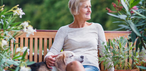 Older woman sitting on a wooden bench outdoors with a dog resting on her lap, surrounded by plants.