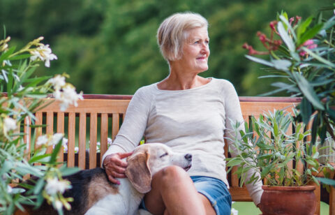 Older woman sitting on a wooden bench outdoors with a dog resting on her lap, surrounded by plants.