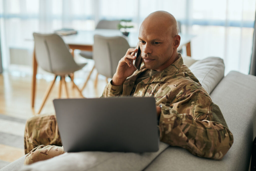 Man in military uniform sitting on a couch, talking on the phone while working on a laptop.