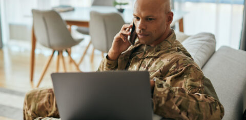 Man in military uniform sitting on a couch, talking on the phone while working on a laptop.
