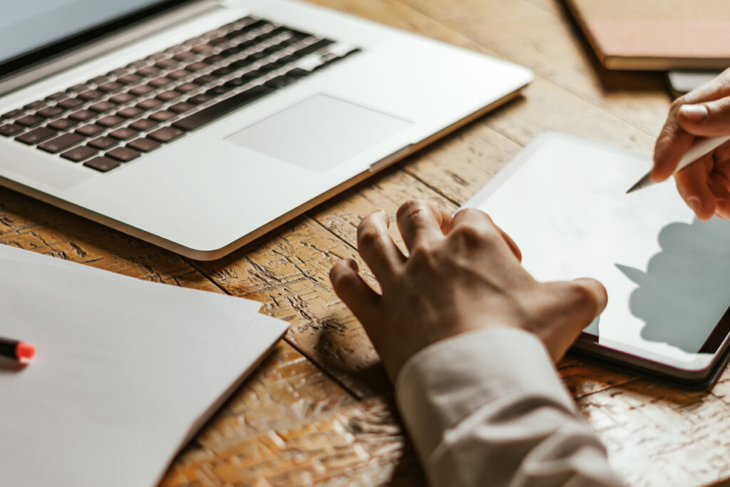 Close-up of a person using a stylus on a tablet next to an open laptop on a wooden desk.