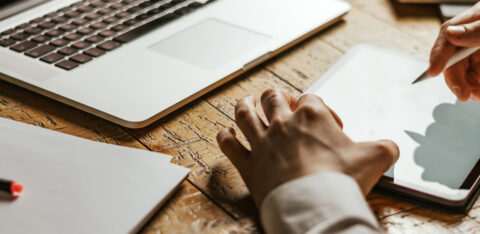 Close-up of a person using a stylus on a tablet next to an open laptop on a wooden desk.