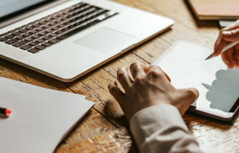 Close-up of a person using a stylus on a tablet next to an open laptop on a wooden desk.