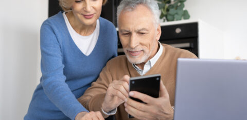 Older couple reviewing information on a smartphone, with papers and a laptop on the table.
