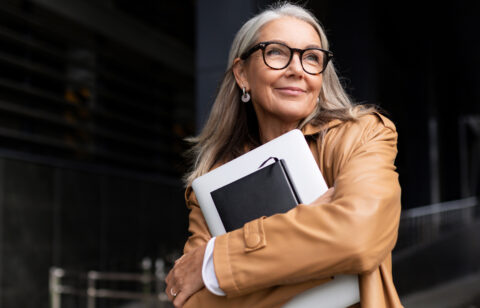Professional-looking senior woman in glasses and a tan coat, carrying work materials and gazing into the distance.