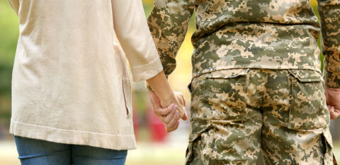 Military service member and civilian partner holding hands outdoors, shown from behind.