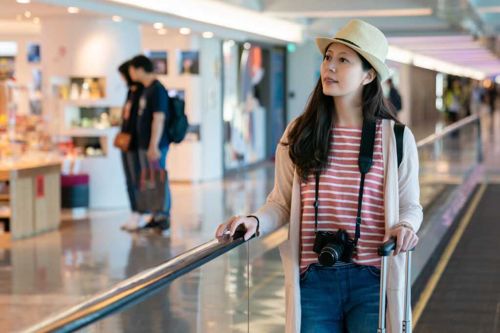 Woman in a striped shirt and straw hat pulling a suitcase through an airport terminal with a camera around her neck.
